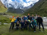 Trekking group posing with poles on green grass beside a river, with the snow-capped Salkantay massif behind