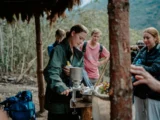 Traveler grinding coffee beans with a hand mill under a thatched shelter while the trekking group watches
