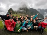 Salkantay Trek group in rain ponchos cheering with arms raised in front of a cloud-covered Machu Picchu