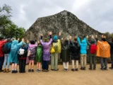 Group of visitors in colorful rain ponchos standing with hands raised in front of the Sacred Rock of Machu Picchu