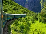 View from a train window of blue carriages curving along the tracks through a green canyon towards Aguas Calientes