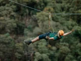 Traveler in an orange helmet flying superman-style along a zipline cable above dense green cloud forest